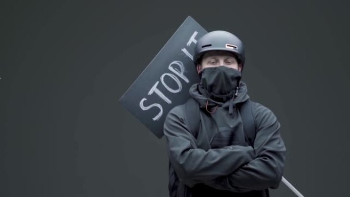 穿着黑色面具和带有纸板标志的头盔的男性抗议者在灰色工作室背景上处于防御位置