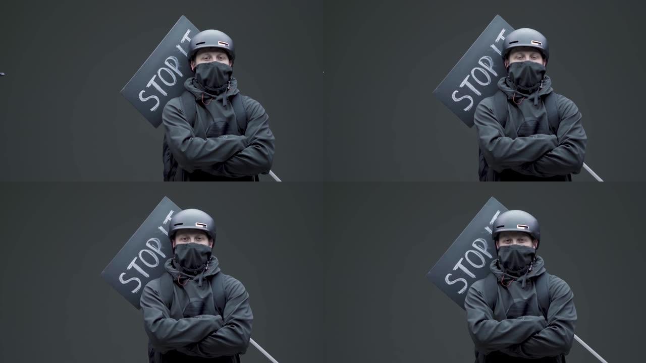 穿着黑色面具和带有纸板标志的头盔的男性抗议者在灰色工作室背景上处于防御位置