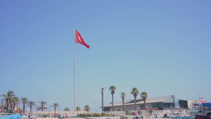 红色土耳其国旗在带有渔船的城市码头下的湛蓝天空中挥舞。