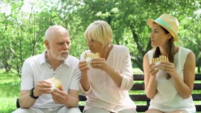 年长者和十几岁的女儿坐在长凳上，在公园里吃火腿烤三明治