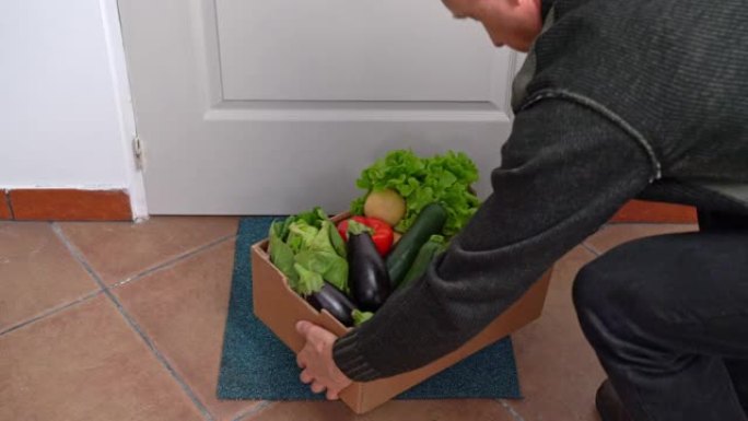 在检疫和大流行期间，一名送货员将一盒新鲜蔬菜放在门前的地板上。食品供应概念。