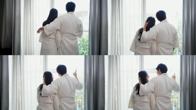 穿着浴袍的亚洲夫妇早上在豪华酒店卧室醒来时打开窗户上的窗帘