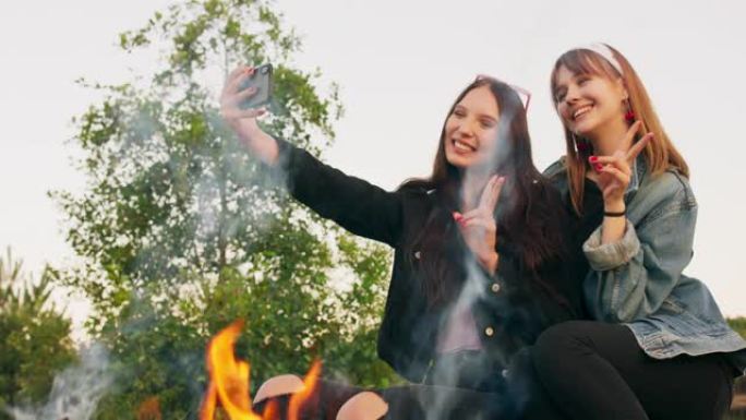 迷人的多种族两个年轻女孩在树林里燃烧的野营篝火旁自拍，微笑着