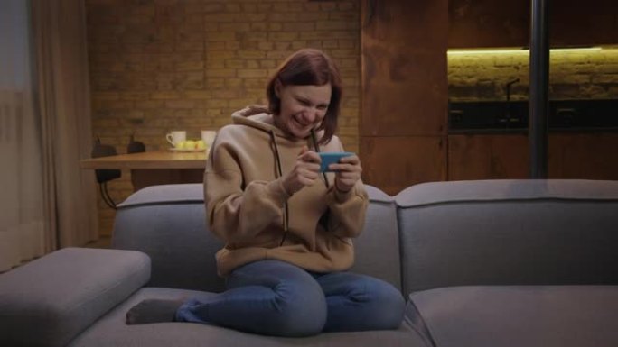 30多岁的女人在手机上玩手机游戏，坐在沙发上赢得游戏。女性一边玩手机一边笑。