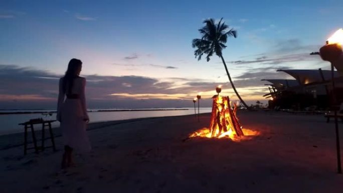 假日时，穿着漂亮衣服的模特女孩的篝火剪影在海滩浪漫的夜晚度假酒店散步。日落时的热带岛屿豪华海滩别墅。