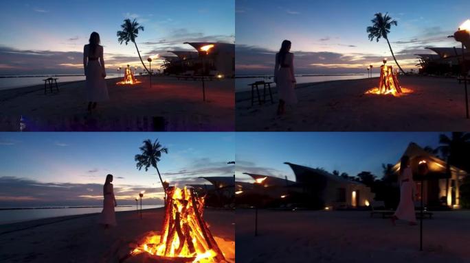 假日时，穿着漂亮衣服的模特女孩的篝火剪影在海滩浪漫的夜晚度假酒店散步。日落时的热带岛屿豪华海滩别墅。