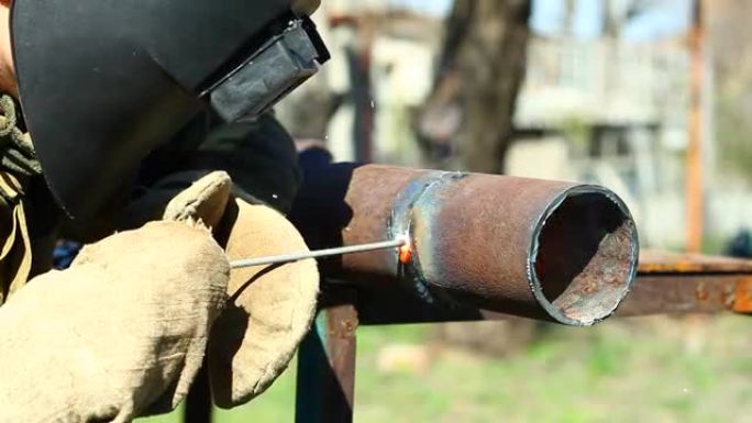 在阳光明媚的天气下，穿着防护服和手套的男子焊工在室外的铁制工作台上焊接带有电极的铁制厚管。特写。