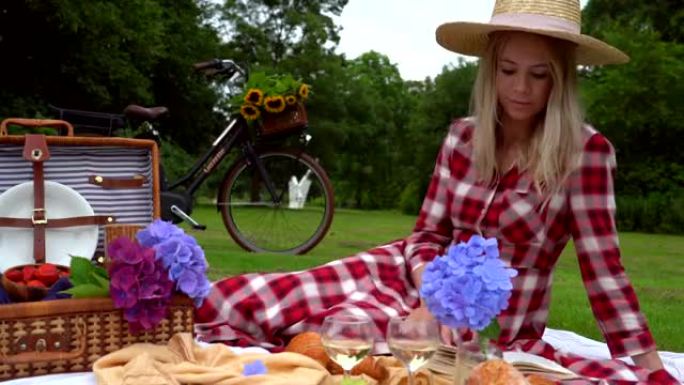 穿着红色方格连衣裙和帽子的女孩坐在白色针织野餐毯上看书，喝酒。阳光明媚的夏天野餐，有面包、水果、花束