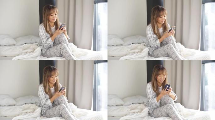 穿着时尚家居服的迷人亚洲女性正坐在床上，手里拿着智能手机