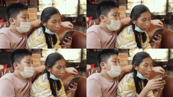 戴着口罩的亚洲男孩看着他的妹妹在智能手机上观看病毒视频，同时在餐厅放松，生活方式概念。