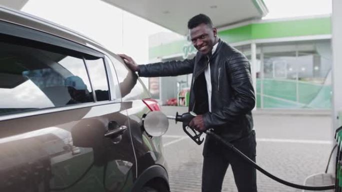 非裔美国时尚男子在他吉普车附近的加油站。该名男子为他的汽车加油，看着相机微笑。燃料，加油站，汽油价格