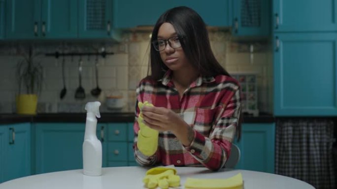 年轻漂亮的非洲裔美国妇女厌倦了家政服务，脱下坐在蓝色厨房餐桌上的黄色橡胶手套。