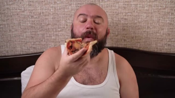 一个大胡子的男人坐在家里的床上，通过网络摄像头进行视频通话，然后吃披萨。用满嘴交流