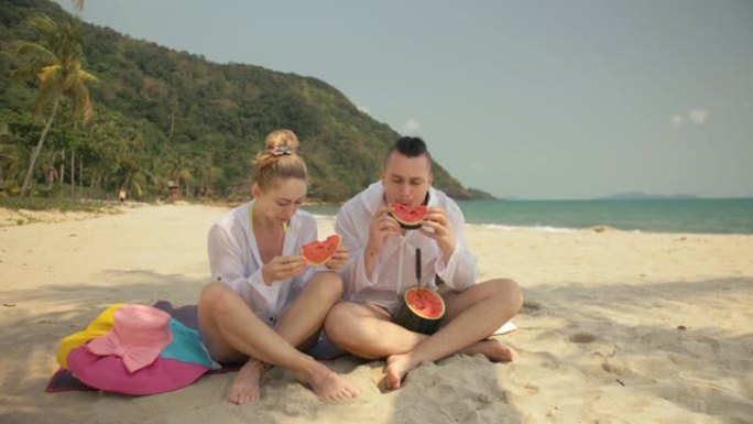 欢快的爱情夫妇在热带沙滩上拿着和吃西瓜片。浪漫的恋人两个人度过夏季周末。
