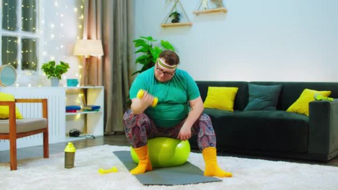 有一个肥胖的男人坐在一个明亮的黄色健身球上，他正在用黄色的粪便铃铛在手臂上工作，他看起来非常集中