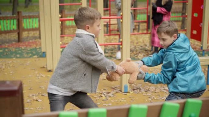 在秋季公园的操场上，高加索男孩为玩具而战。两兄弟在户外拉泰迪熊。冲突与童年观念