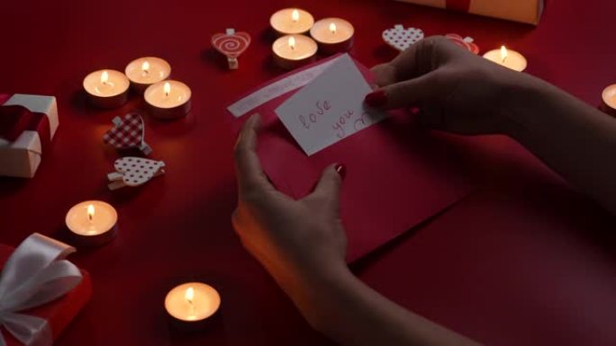 女人的手打开信封，拿出一张写着爱你的情人卡。红色桌子的俯视图，带有燃烧的蜡烛，装饰性的心形和礼物。情