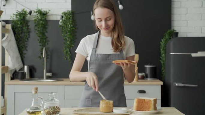 一个女人站在厨房的围裙里，在烤面包上撒上花生酱