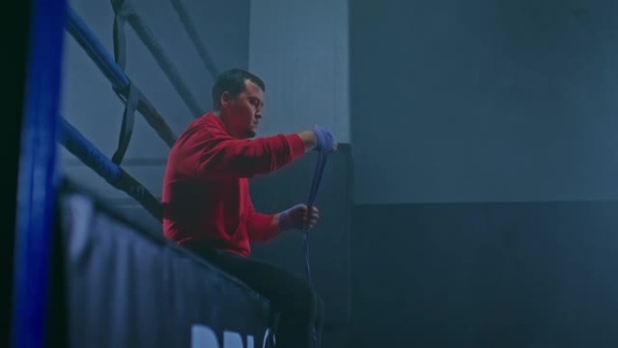 一名男性拳击手坐在烟雾弥漫的黑暗健身房里的拳击台上，手上缠着保护性绷带。战士准备战斗，双手和手指都裹