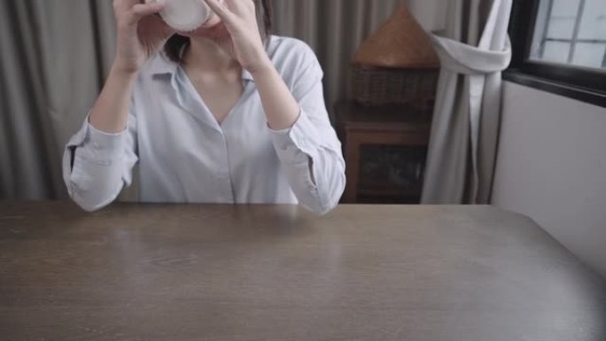 亚洲女性喝热饮料，拿着茶杯坐在办公桌上喝酒，早上喝咖啡，自己呆在家里，一天开始，咖啡因成瘾的习惯，平