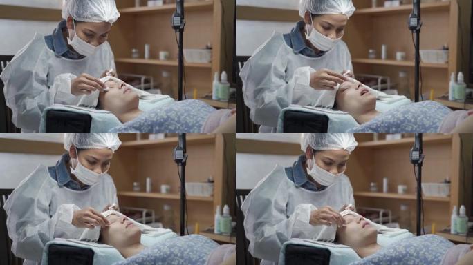 亚洲化妆师制作睫毛沙龙程序。女人的睫毛延伸。专业佩戴医用口罩帽子和个人防护用品套房，新型冠状病毒肺炎