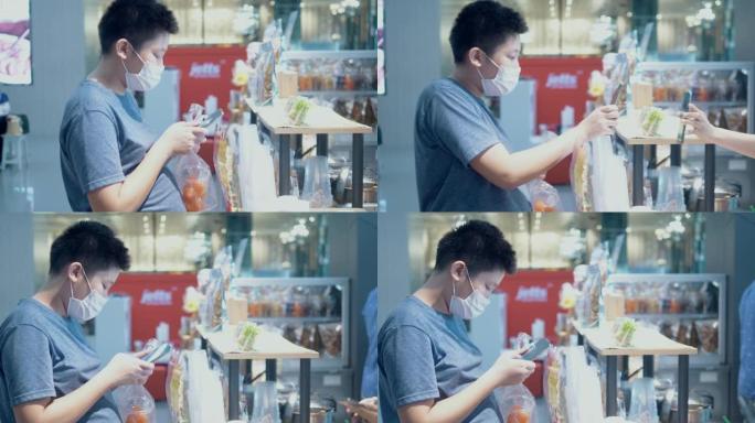 戴着口罩的亚洲男孩购买一些零食和饮料，然后在美食广场通过手机二维码付款，这是新的正常生活方式概念。
