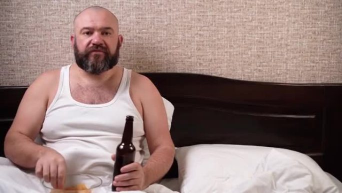 一个丰满的大胡子男人看电视，喝啤酒，吃薯条，坐在家里的床上。复制空间