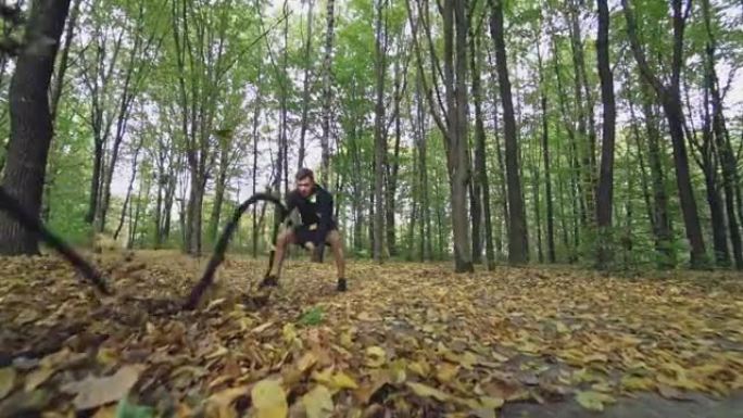 运动员在秋天的森林里用战斗绳波浪。穿着黑色运动服的运动员正在用绳子训练，然后离开。慢动作。