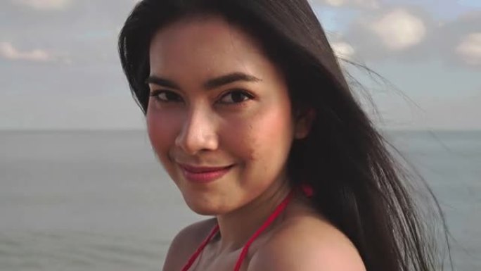 穿着比基尼的年轻亚洲美女在镜头前微笑的肖像。夏天天气下，热带海滩上的头发随风吹来。旅行和生活方式概念