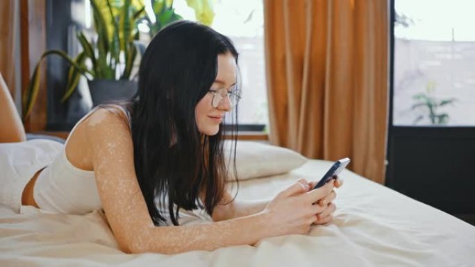 色素沉着不均匀的年轻女子躺在床上，在智能手机上聊天，慢动作