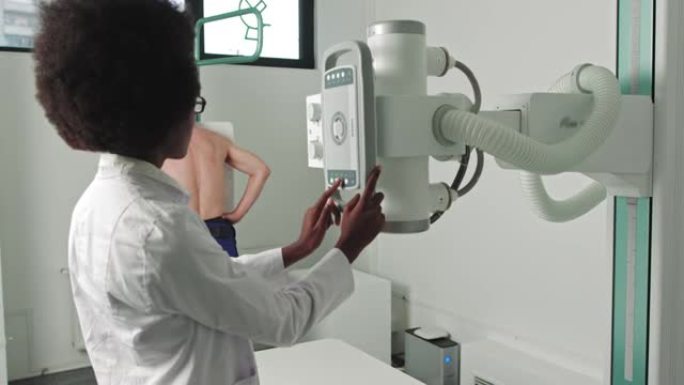 在医院里，一名男子面对墙壁站着，而医疗技术人员则调整x光机进行扫描。扫描骨折，四肢骨折，胸部，癌症或