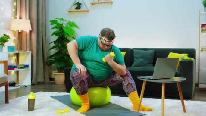 有一个戴着眼镜的肥胖男人，他留着胡须，坐在黄色的瑜伽球上，他非常担心用粪铃锻炼，他正在看着笔记本电脑