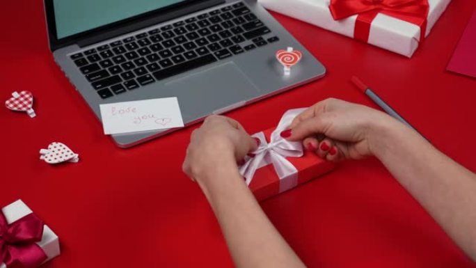 女人的手正在包装礼品盒并系白色蝴蝶结。带笔记本电脑、礼物、心形和情人节的红色桌子的俯视图。为恋人假期