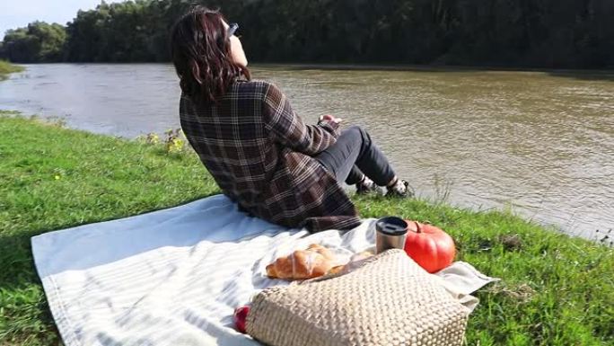 年轻漂亮的女人坐在毯子上拿着书。河边野餐