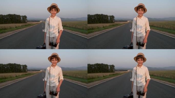 夏天看着相机的乡村道路上带着复古手提箱微笑的小快乐男孩的肖像