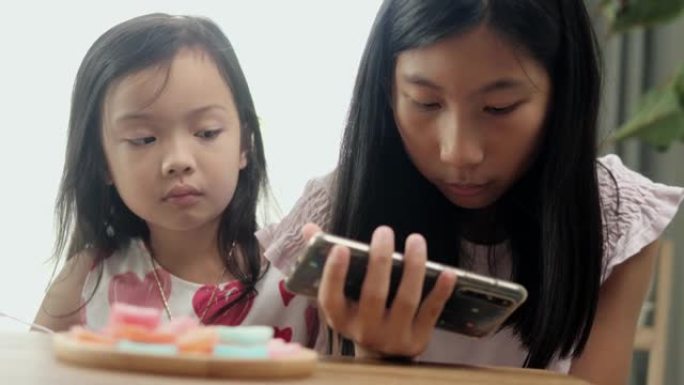 快乐的亚洲女孩通过智能手机观看病毒视频，并在家里一起吃一些甜蜜的零食，生活方式概念。