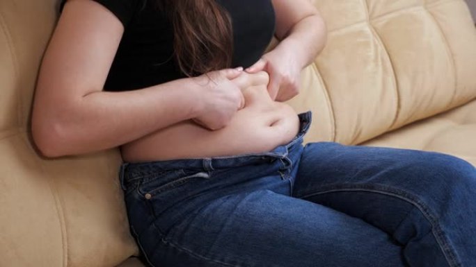 穿着牛仔裤的无法辨认的女人坐在沙发上时会感觉到腹部的褶皱