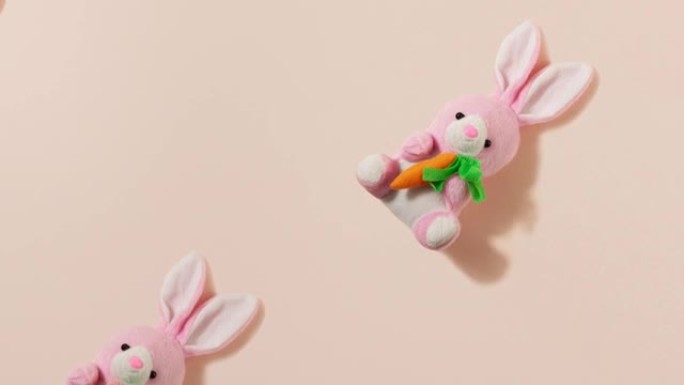 慢动作。粉色毛绒小复活节兔子，粉色米色背景上有红色胡萝卜，玩具兔子作为图案。复活节假期概念。复活节背