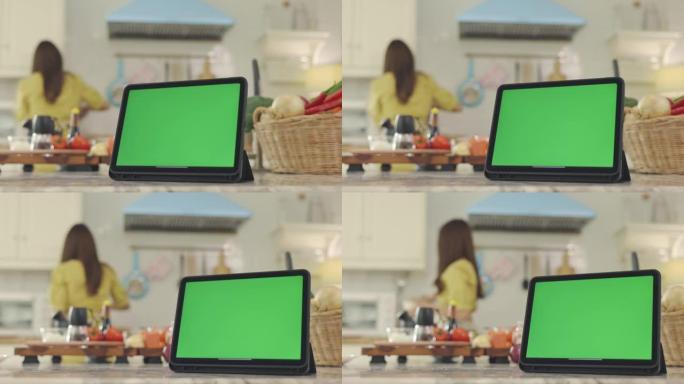 平板电脑带有绿色屏幕，背景优美，在屋子里做饭，带着幸福的脸，妻子在家里开心地做早餐。