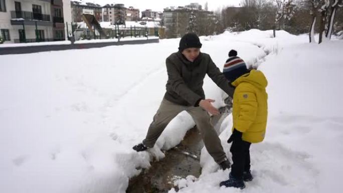 年轻的父亲在雪中帮助儿子越过溪流