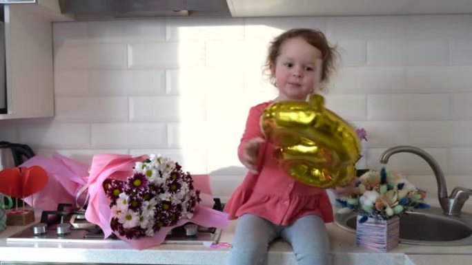 可爱的有趣的小女孩拿着金色气球8 8坐在厨房的桌子上，手里拿着一束春花，在家问候母亲或祖母。母亲节。