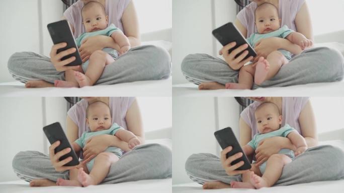 快乐微笑的亚洲年轻母亲在家里拥抱刚出生的婴儿时使用手机。