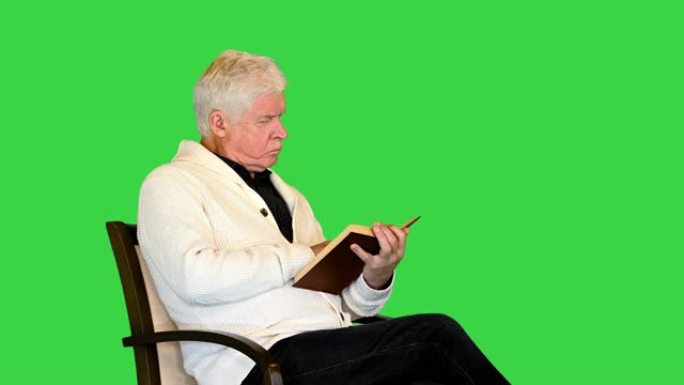 高年级男子坐在绿色屏幕上的椅子上学习一本书，色度键
