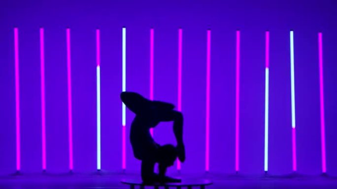 体操运动员的剪影在多色霓虹灯的背景下在工作室里进行杂技练习。女孩做倒立和胸立，然后坐在裂口上。特写
