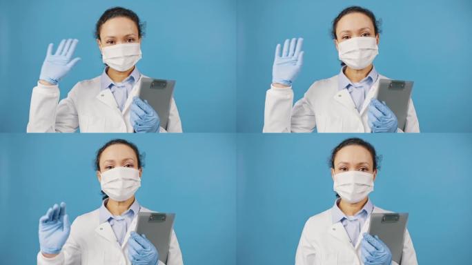 友好的女医生戴着防护口罩，拿着剪贴板向相机挥手致意，蓝色背景