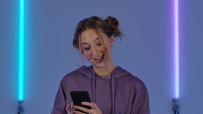 年轻迷人女人的肖像正在手机上发短信，欢欣鼓舞。穿着紫色连帽衫的时装模特在黑暗工作室明亮的霓虹灯背景下