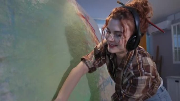 创意氛围，戴着耳机的才华横溢的女孩艺术家用手在车间的画布上蘸上生动的颜料，在运动中的相机