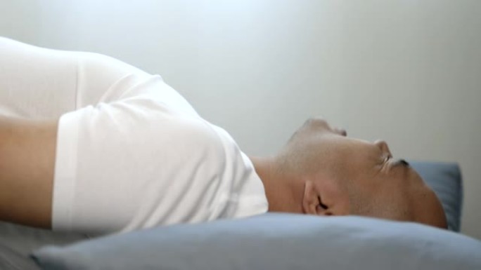 穿着白色t恤的30岁左右的亚洲秃头男人睡不好觉。他有头痛、失眠和噩梦。