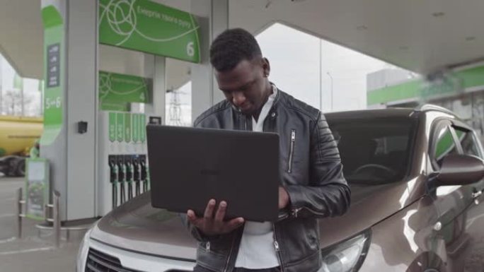 一名非裔美国自由职业者手里拿着一台笔记本电脑，靠在一辆汽车上。工作自由职业者。加油站的车。燃料，汽油