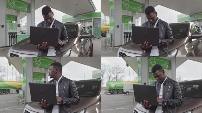 一名非裔美国自由职业者手里拿着一台笔记本电脑，靠在一辆汽车上。工作自由职业者。加油站的车。燃料，汽油